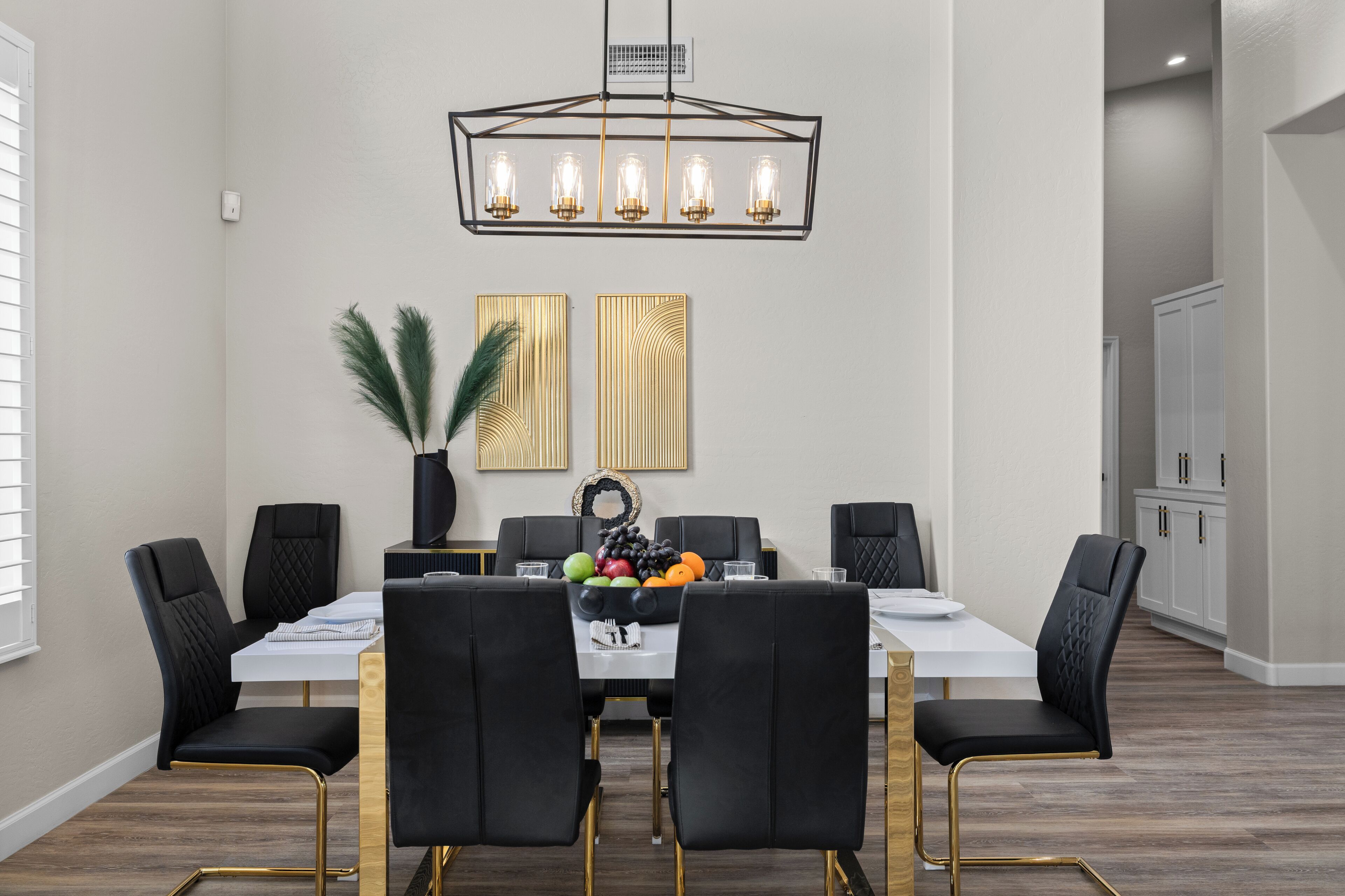 Un comedor con una mesa rectangular, sillas tapizadas en negro, una araña y dos paneles decorativos en la pared.