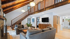 Ein geräumiger Wohnbereich mit einem großen, grauen Ecksofa, einem Kamin und einer Holztreppe, die zu einer oberen Ebene führt.