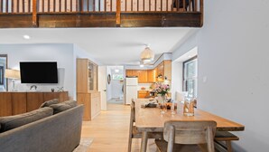 Ein modernes Wohnzimmer mit einer Couch, einem Holzespeisetisch und Stühlen. Im Hintergrund befindet sich eine Küche mit Holzschränken und einem Kühlschrank.