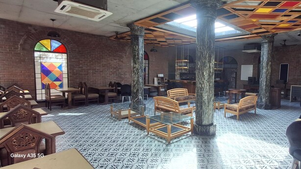 A spacious indoor area with high ceilings, exposed brick walls, and a patterned floor. There are multiple seating areas with tables and chairs, and a large stained glass window is visible. - Galleria Hotel Ajmer (Ajmer)