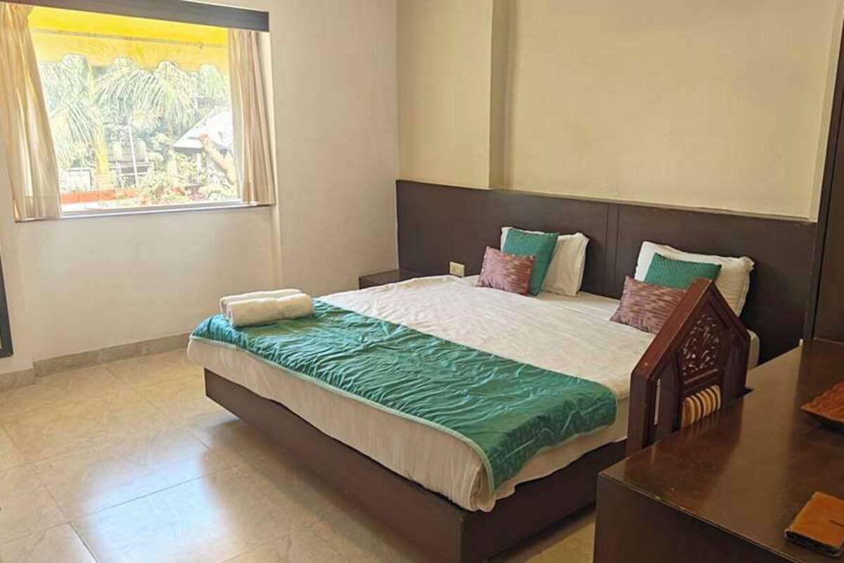 A hotel room with a bed, a nightstand, a window with curtains, and a view of greenery outside.