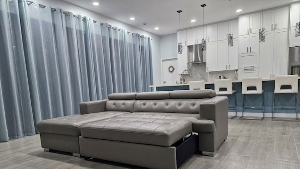 Una sala de estar moderna con un sofá grande, una zona de cocina con armarios blancos y una cortina gris.