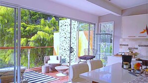 A modern kitchen with white cabinets and a large island, a dining area with wicker furniture, and a sliding glass door leading to a deck with outdoor seating. - Grand Haven with Sea view! For Executives and Remote! Adults only! City Close! (Castlecrag)