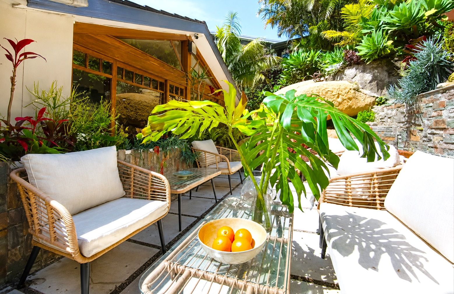 Un espace extérieur aménagé avec des meubles en osier et une table en verre, entouré d’une végétation tropicale luxuriante.