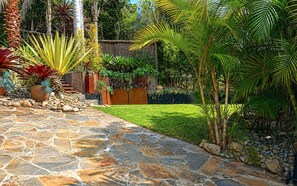 Un sentier de pierres mène à une structure en bois, elle-même entourée d’une végétation luxuriante et de diverses plantes.