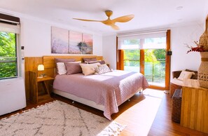 Une chambre à coucher avec un lit, un ventilateur de plafond, une grande fenêtre et une vue sur la verdure.