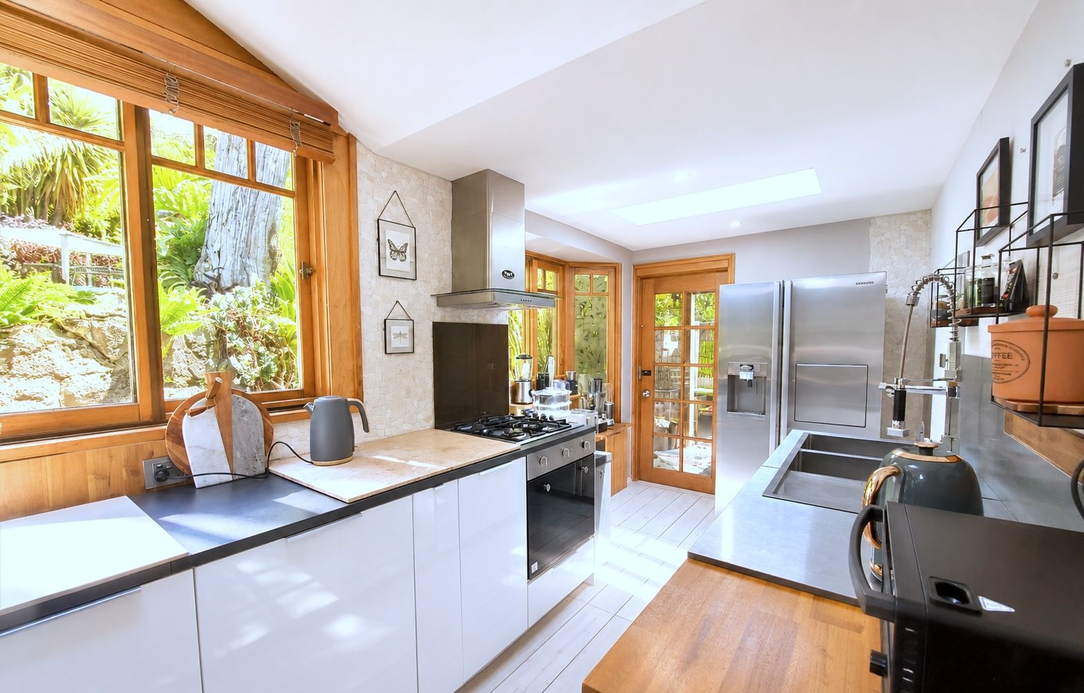 Une cuisine moderne dotée d’un îlot central, d’appareils électroménagers en acier inoxydable et offrant une vue sur le jardin grâce à de grandes fenêtres.