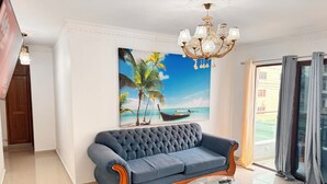 Een woonkamer met een blauwe bank, een glazen salontafel, een kroonluchter en een schilderij van een strandgezicht.