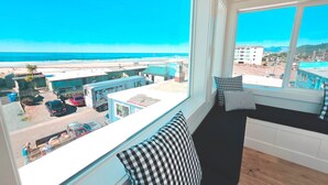 Ein Zimmer mit großem Fenster mit Blick auf einen Strand und eine Stadtlandschaft. Auf einer Bank liegt ein schwarz-weiß kariertes Kissen und ein weiteres Kissen mit Streifenmuster.