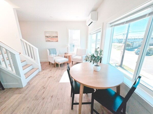 A bright, open living area with a wooden staircase, a round dining table with chairs, a potted plant, and large windows offering a view of the outside.