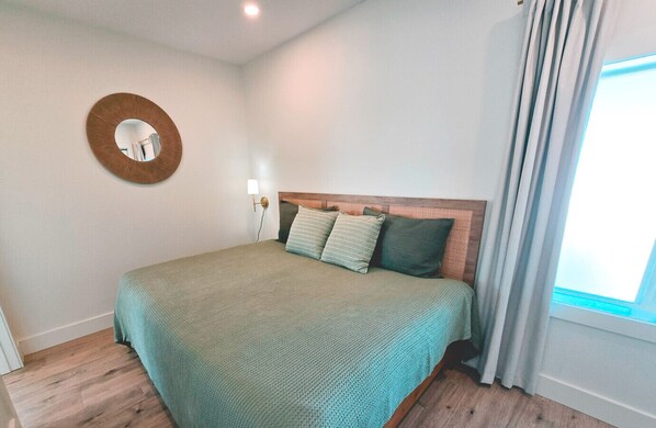 A neatly arranged bedroom with a bed, a wall-mounted mirror, and a window with curtains.