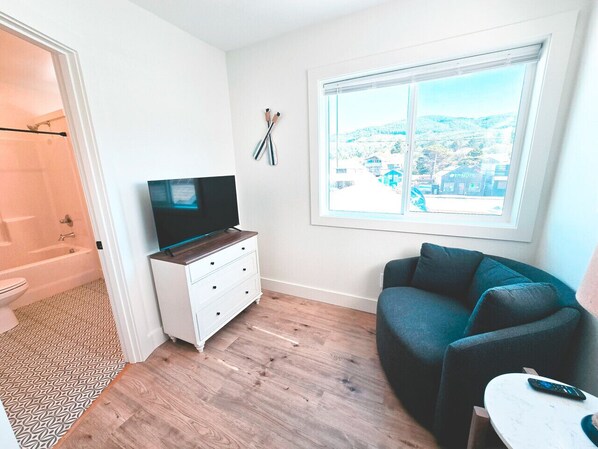 Una sala moderna con un televisor sobre un aparador blanco, un sofá azul oscuro y una ventana con vista a la ciudad.
