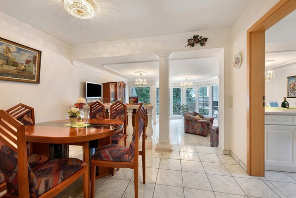 A dining area with a round table and chairs, a kitchen with white cabinets, and a view of the outdoors through large windows. - Apartment 'Linas Lieblingsplatz' with Balcony & Piano (Uhldingen-Mühlhofen)