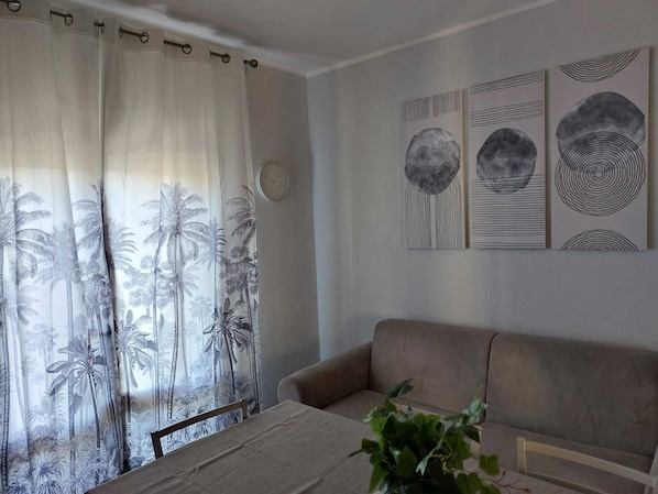 A living room with a beige sofa, a bed with a white bedspread, and a window with palm tree curtains. There are three wall art pieces above the sofa. - Accommodation With Terrace and Parking (Rosolina)