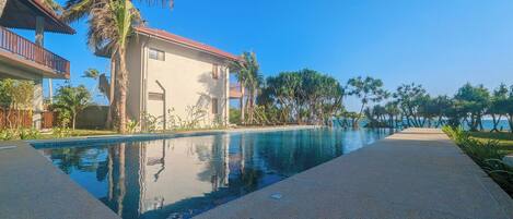 A swimming pool with a clear view of the sea, surrounded by palm trees and a building with a red roof.