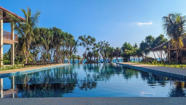 A swimming pool with clear blue water, surrounded by palm trees and a building with a red roof. - Arinna Beach Resort (Tangalle)