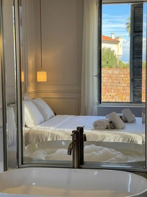 A hotel room with a bed, a large mirror, a window with shutters, and a view of a building outside. - Lily (Cannes)