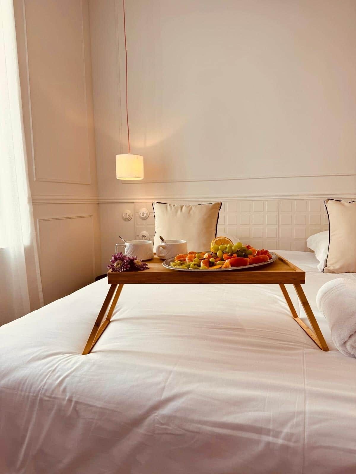 Een hotelkamer met een net opgemaakt bed, een houten nachtkastje met een fruitplateau, een lamp en een vaas met bloemen.