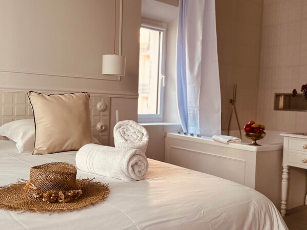 A hotel room with a neatly made bed, a bedside table with a bowl of fruit, and a window with a curtain. - Lily (Cannes)