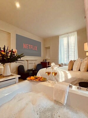 A hotel room with a bed, a coffee table with a fruit platter and glasses, a bathtub with bubbles, and a wall with a Netflix sign. - Lily (Cannes)