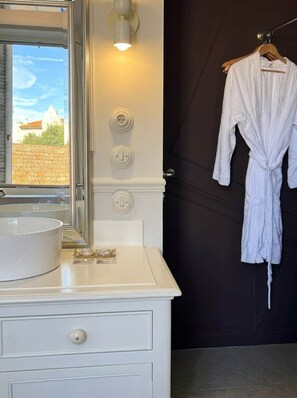 A bathroom with a white vanity, a round mirror, a towel rack with a white robe, and a wall-mounted light fixture. - Lily (Cannes)