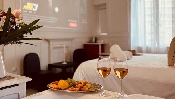 A hotel room with a bed, a dining table with a fruit platter and two glasses of wine, a vase with flowers, and a projector screen displaying a movie. - Maison LYO - "Lys" Romantic Luxury Suite (Cannes)