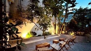 Un área de estar al aire libre con mobiliario de madera y piso de grava, rodeada de follaje y una fachada de edificio.