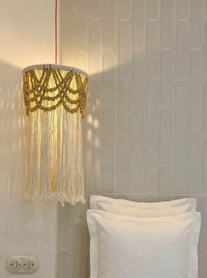 Una habitación con pared blanca, una cama blanca con almohadas y una lámpara colgante con flecos dorados.