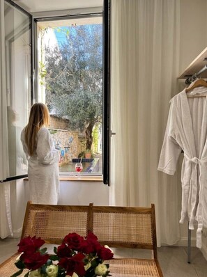 Ein Zimmer mit großem Fenster, einer Korbsitzbank, einem Holztisch mit Blumen und einem weißen Bademantel an einem Gestell.