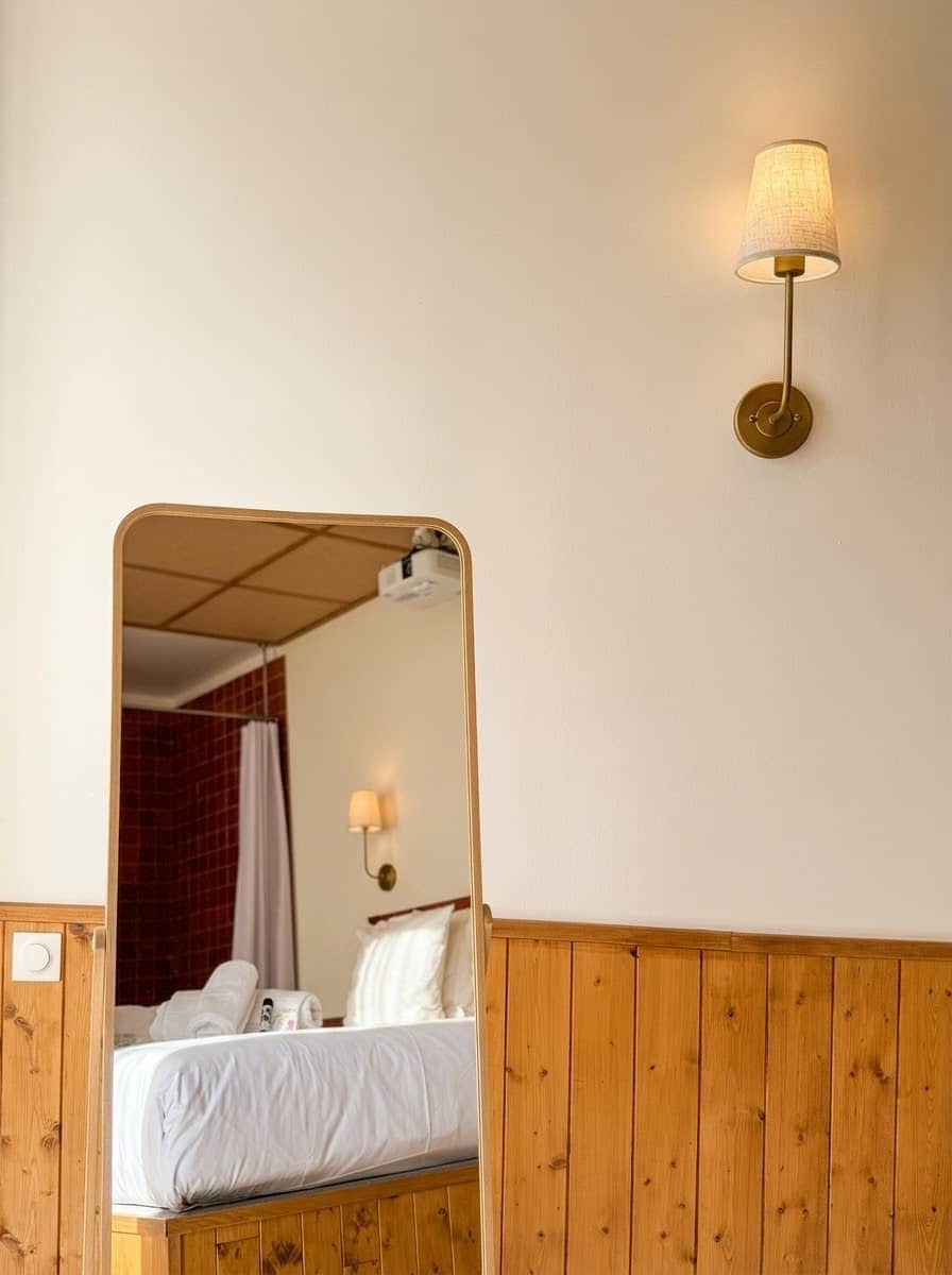 A hotel room with a wooden panel wall, a bed with white bedding, a wall-mounted lamp, and a mirror reflecting the room.
