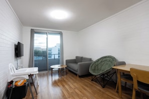 Ein kleiner, moderner Raum mit einem grauen Sofa, einem grünen Klappsessel, einem Holztisch und einem weißen Fernseher, der an der Wand befestigt ist.