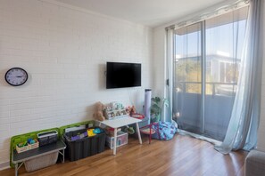 A living room with a white brick wall, a mounted TV, a wooden floor, and a sliding glass door leading to an outdoor area. - Cosy 2 BDR APT in heart of Fremantle with Parking (Fremantle)