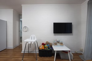 Ein Zimmer mit weißer Ziegelwand, einem weißen Stuhl, einem kleinen Tisch mit Spielzeug und einem an der Wand befestigten Fernseher.