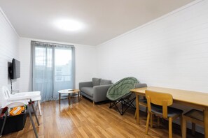 A compact living space with a sofa, a small table, a folding chair, a television mounted on the wall, and a window with curtains. - Cosy 2 BDR APT in heart of Fremantle with Parking (Fremantle)