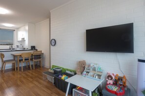Een kamer met een keukenhoek, een eettafel met stoelen, een aan de muur bevestigde televisie en een speelruimte met speelgoed.
