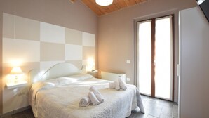 Un dormitorio con cabecera a cuadros, dos lámparas de noche, una cama con toallas enrolladas y un ventanal corredero que da a un balcón.
