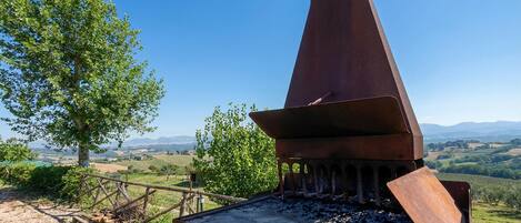 A rustic outdoor grill with a chimney, surrounded by a wooden fence and a tree, overlooking a hilly landscape.