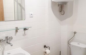 Ein Badezimmer mit weißen Fliesen, einem Glasregal, einem Waschbecken, einer Toilette und einem Spiegel.