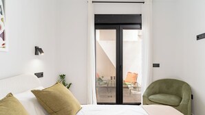 Una camera da letto con un letto, una poltrona verde, una porta-finestra che dà su un balcone con una sedia e un tavolo.