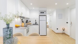 Una cucina moderna con mobili bianchi, un forno a microonde integrato e un frigorifero. Si nota un tavolo da pranzo con sedie e un vaso con fiori.