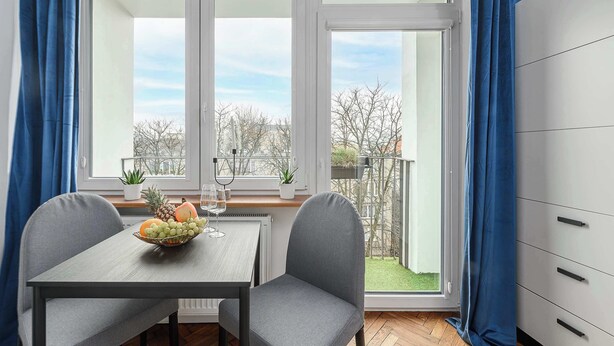 Un coin repas avec une table dressée pour deux personnes, un saladier rempli de fruits et une fenêtre donnant sur un jardin et des bâtiments.