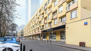 Un immeuble jaune à plusieurs étages, doté de balcons, une devanture de magasin avec une enseigne indiquant « eAZVIMUT.PL », et une voiture garée sur le trottoir.