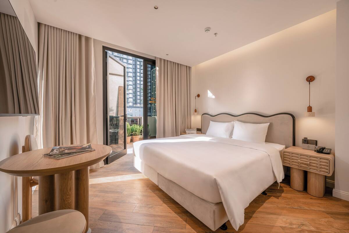 A modern hotel room with a large bed, a wooden side table, a small desk, and a view of the city through the window.
