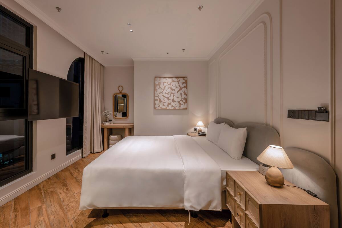 A hotel room with a large bed, bedside table with a lamp, a desk with a chair, and a TV on the left wall.