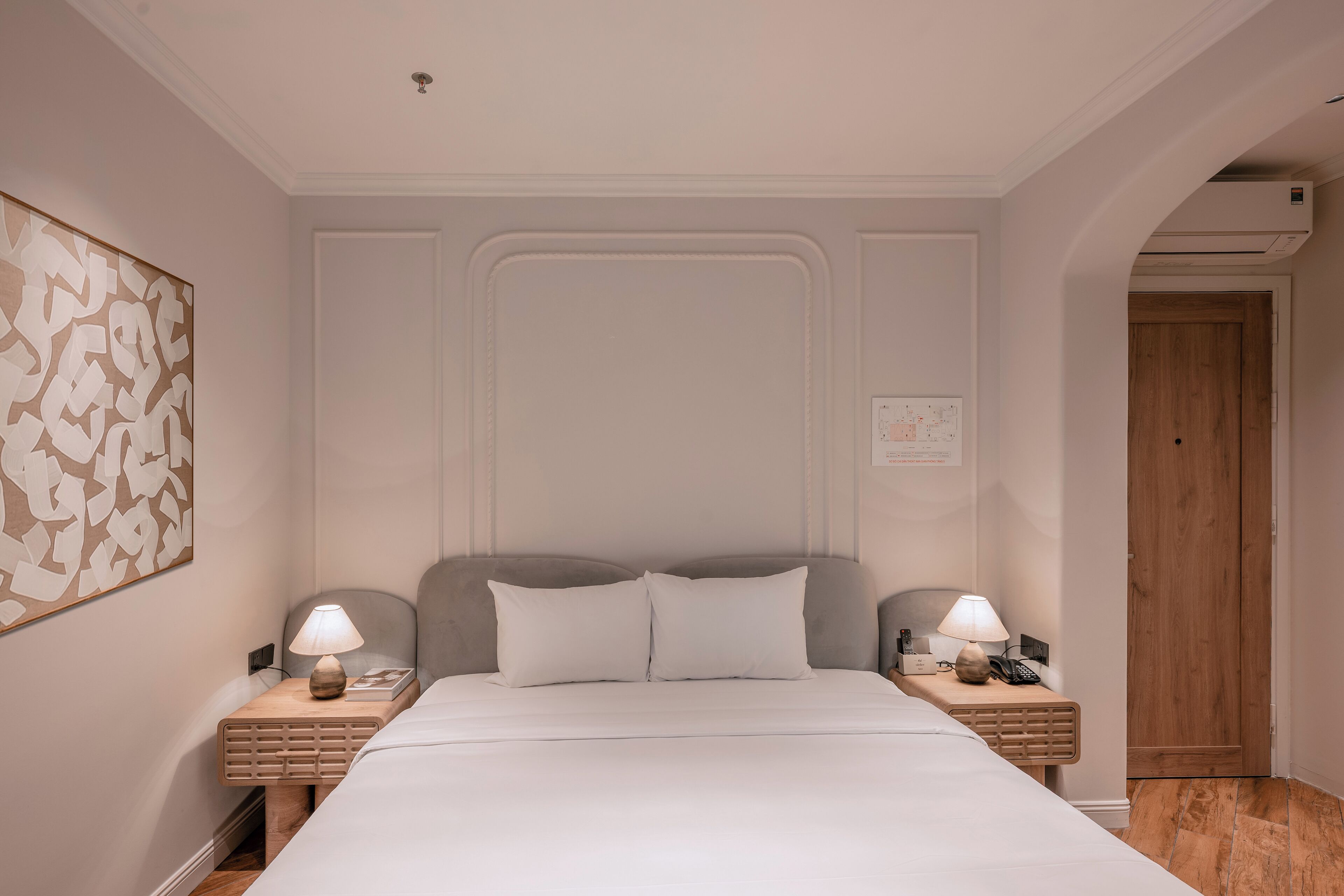 A neatly arranged hotel room with a large bed, two bedside tables with lamps, a wooden door, and a wall-mounted air conditioning unit.