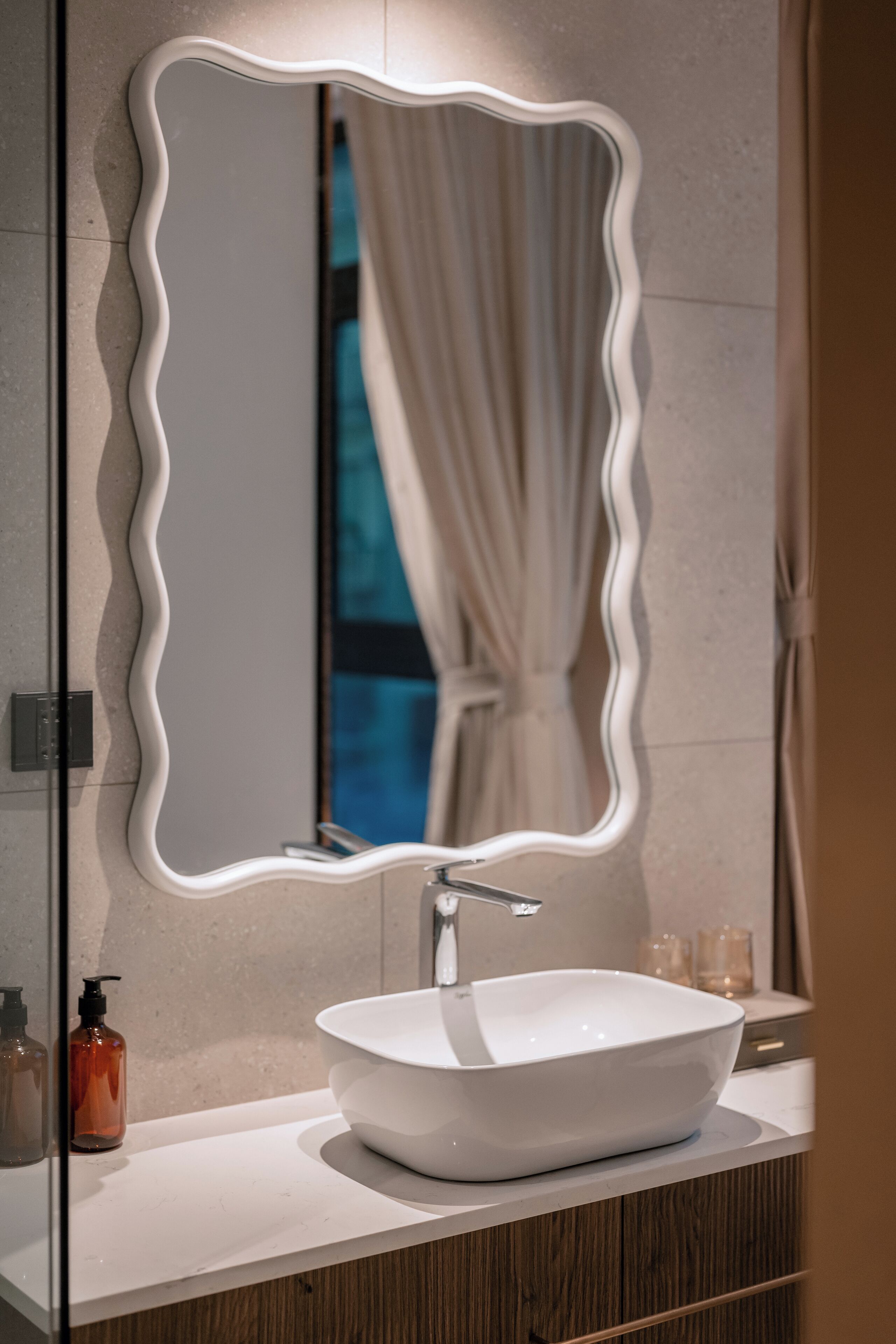 A bathroom with a white vessel sink, a wavy mirror, and a wooden vanity.