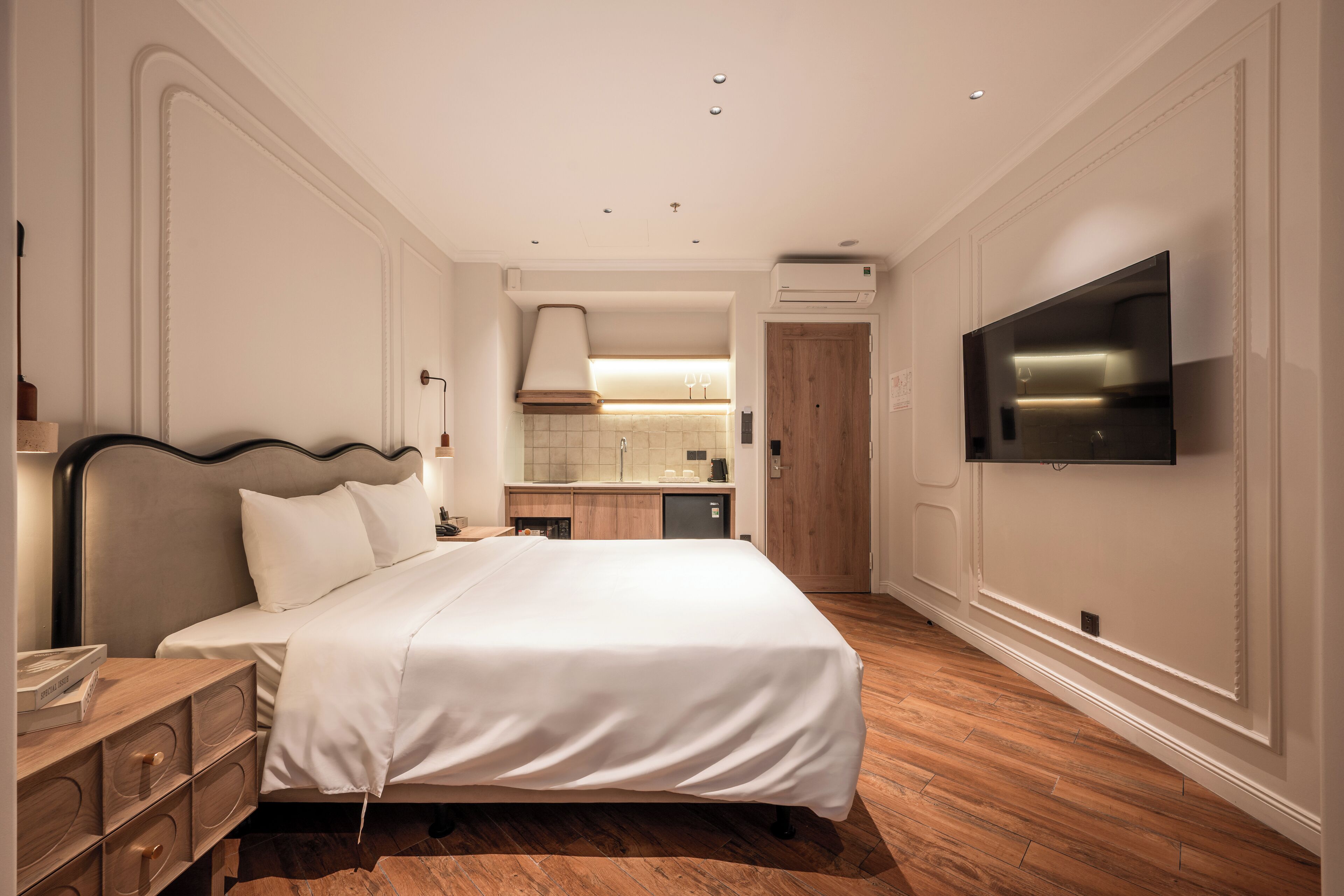 Una habitación de hotel con una cama grande, suelo de madera, un televisor montado en la pared y una kitchenette al fondo.