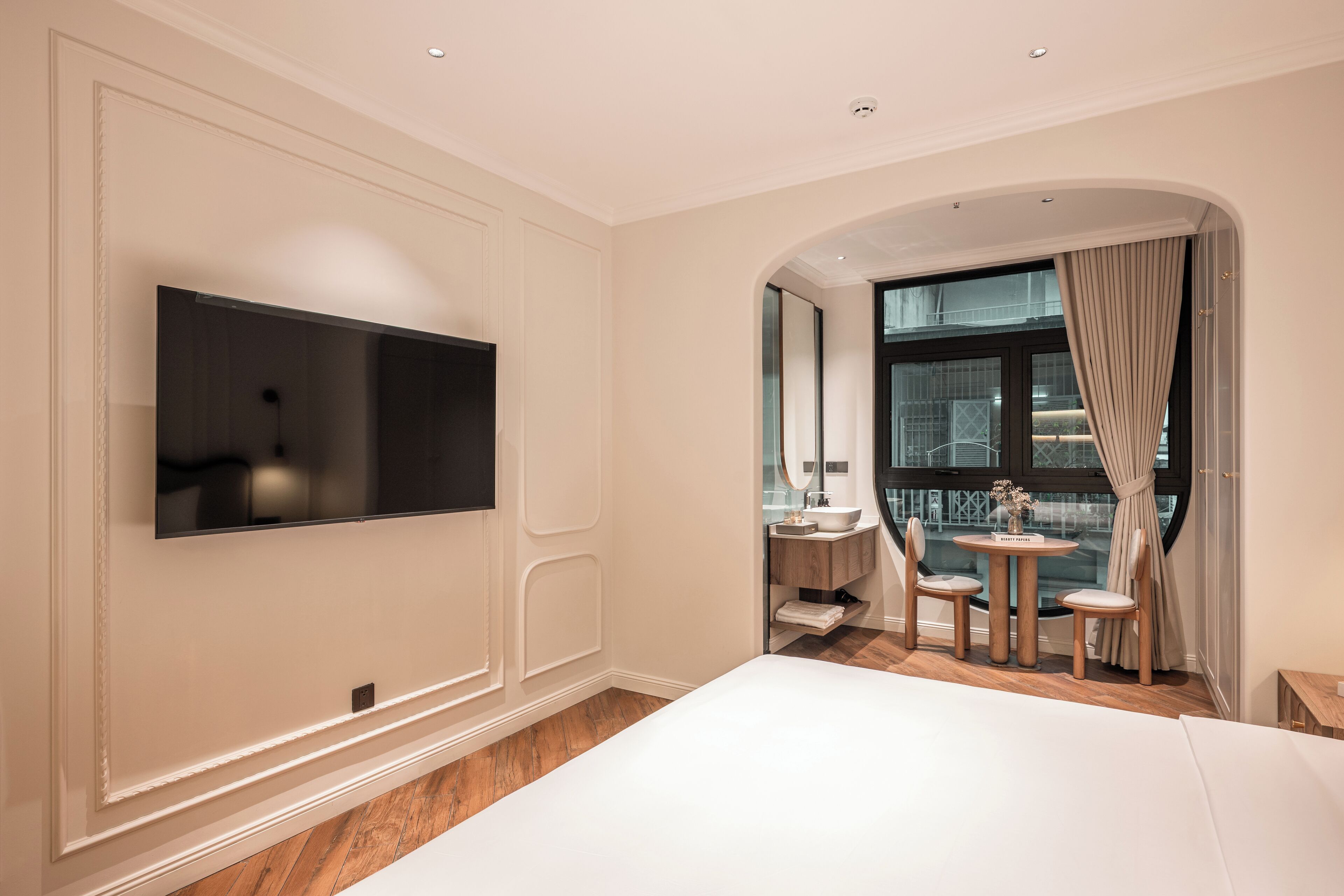 A modern hotel room with a large bed, a flat-screen TV mounted on the wall, a small round table with chairs, a vanity with a mirror, and a view of the city through a large window.