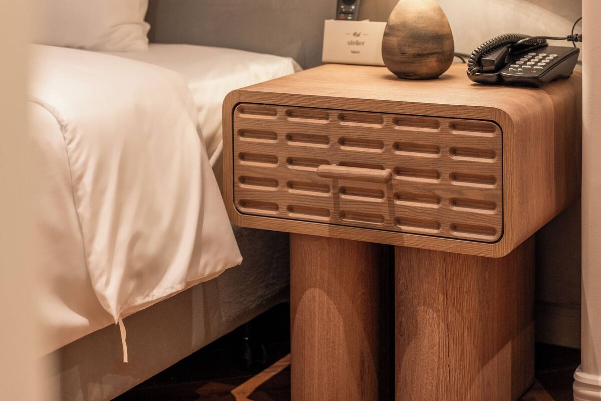A neatly made bed with white linens, a wooden bedside table with a lamp, a telephone, and a small card.