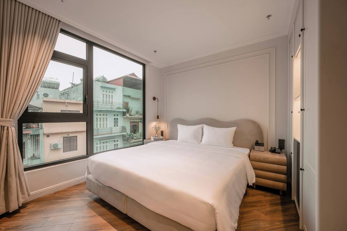 A hotel room with a large bed, a nightstand, a lamp, and a view of buildings outside the window.